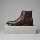 Men's pointed-toe side-zip leather boots in dark brown, straight-on gray pedestal shot